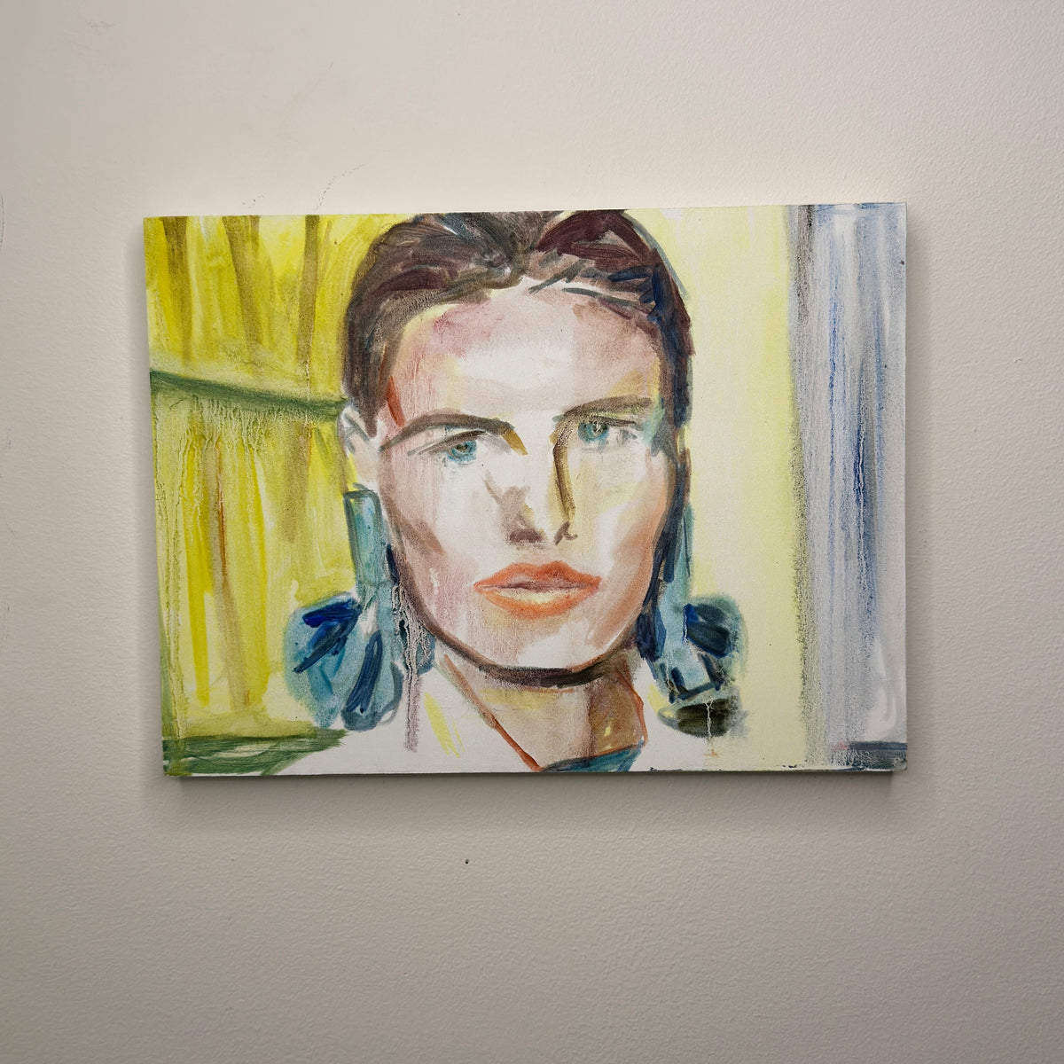 A square canvas painting of a woman's face with blue earrings, displayed on a light gray wall.